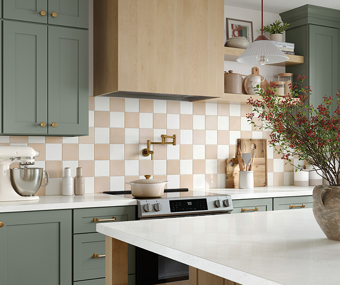 Close-up of modern farmhouse kitchen with sage green cabinets, white quartz countertops, light wood range hood, and beige-and-white checkered tile backsplash.